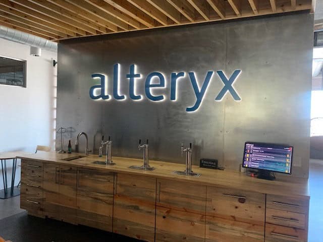 Modern office kitchenette with a large alteryx sign on a metallic wall, wooden cabinetry, beverage taps, a monitor on the counter, and exposed wooden ceiling beams above.