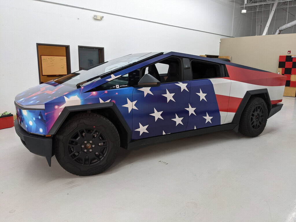 A Tesla Cybertruck with a custom wrap featuring the American flag, displaying blue, white stars on the front half and red, white stripes on the back half, parked indoors on a white floor.