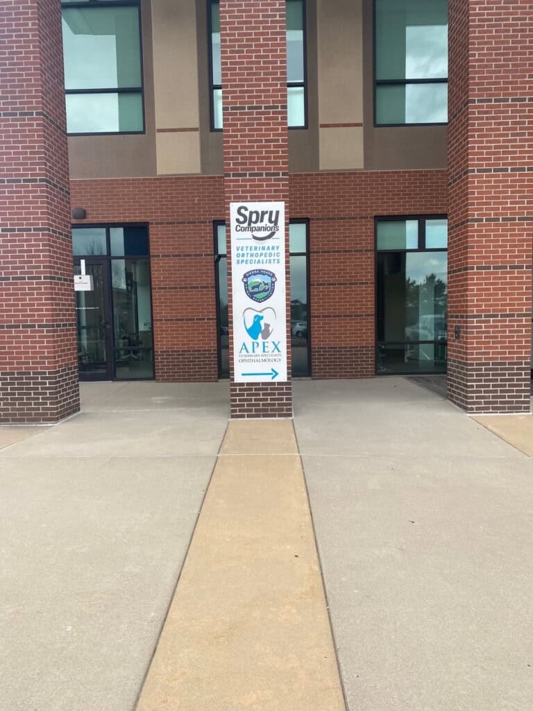 A walkway leads to a brick building entrance with a vertical sign showing logos for Spry Compounding Pharmacy, Veterinary Osteoarthritis Specialists, and Apex Compounding Pharmacy, with an arrow pointing inside.