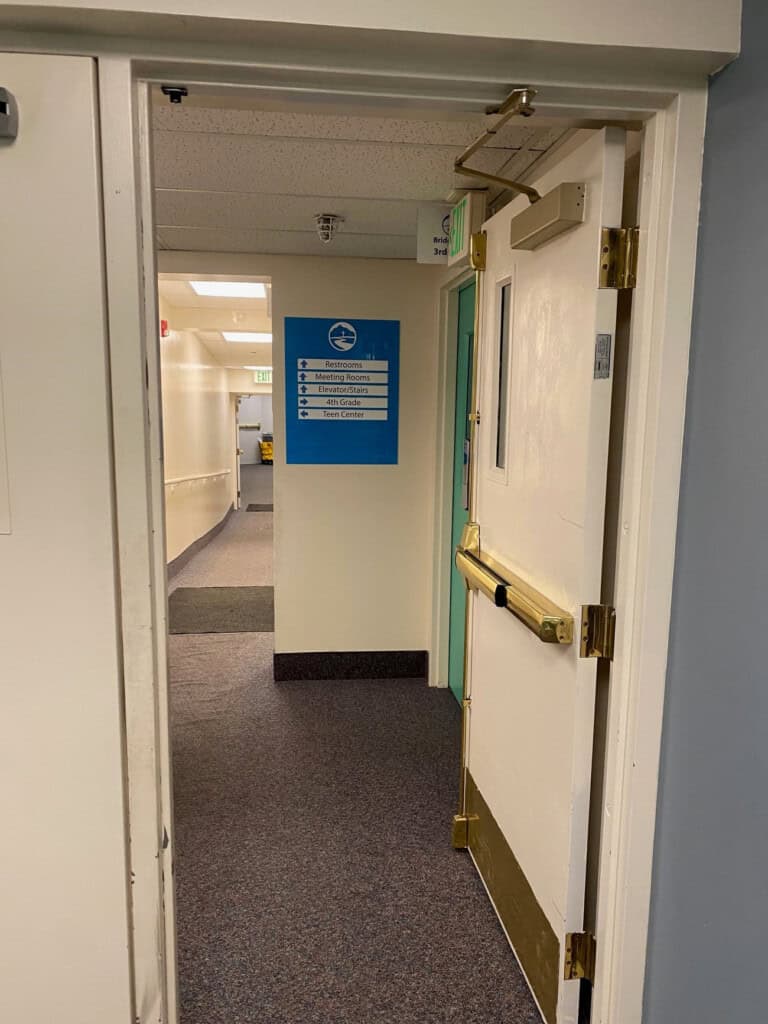 A hallway in a building with an open door. Inside, a sign lists room numbers and destinations, and a yellow cleaning cart is visible at the far end of the corridor.