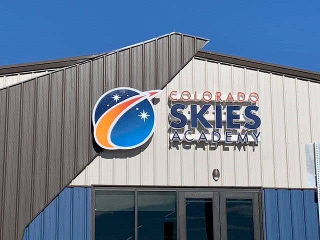 A building exterior featuring a sign that reads “Colorado Skies Academy” with a space-themed logo showing stars and an orange swoosh against a blue background. The sky above is clear and blue.