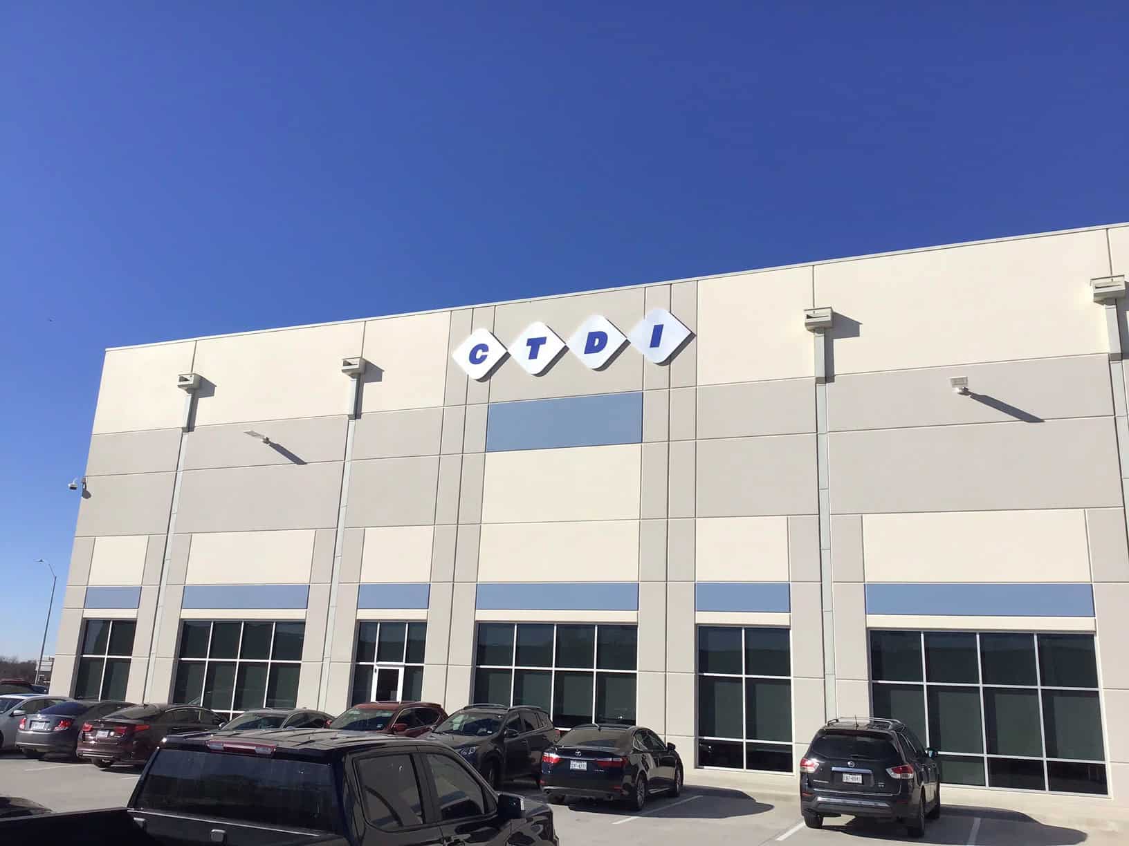 A beige and white commercial building with the letters CTDI displayed in blue on diamond-shaped signs above the entrance; several cars are parked in front under a clear blue sky.