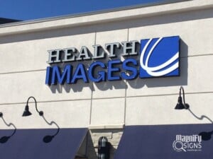 A building sign reads HEALTH IMAGES in bold letters with a blue and white abstract logo to the right. The sign is mounted on a light-colored wall above black awnings.