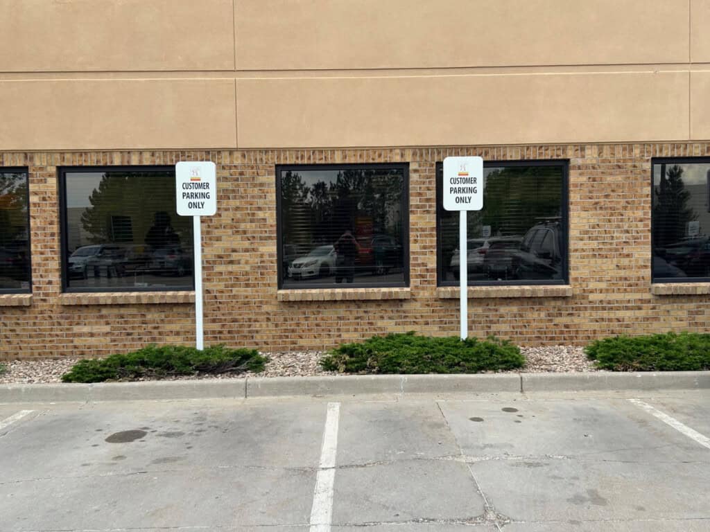 Two empty parking spaces in front of a brick building with large windows. Each space has a sign reading Customer Parking Only. There are shrubs and rocks along the buildings foundation.