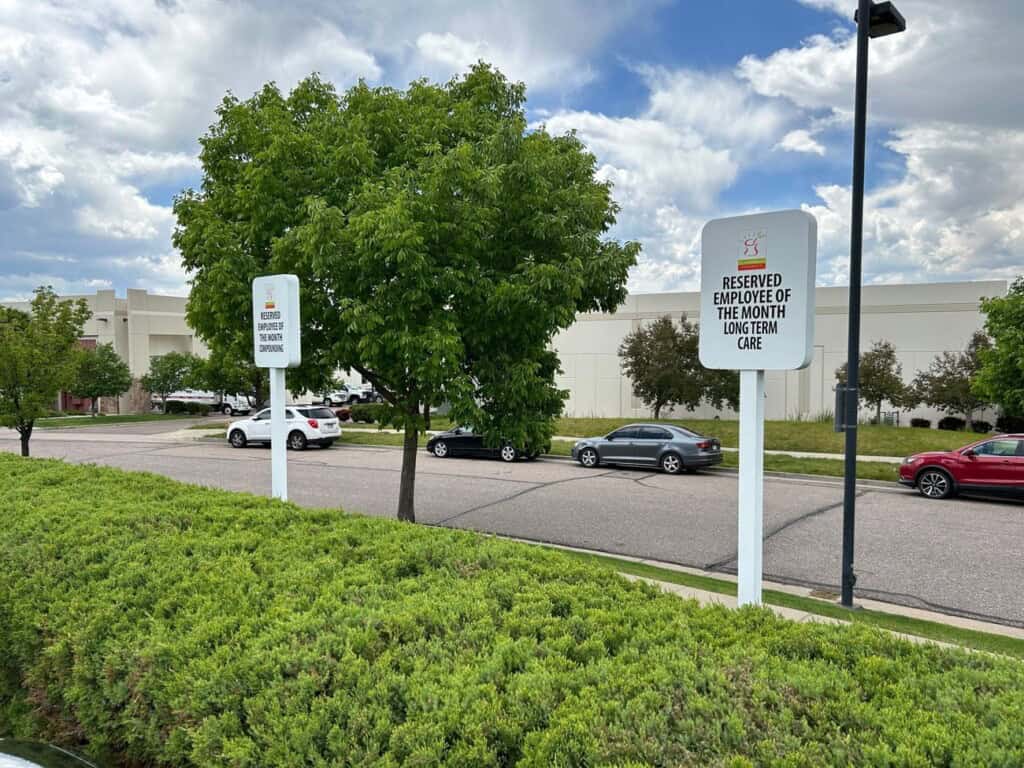 Two parking signs stand next to green bushes. One reads “Reserved Employee of the Month Long Term Care,” while the other reads “Reserved Hospice Nursing Station.” Cars are parked along a curb by a white building with trees nearby.