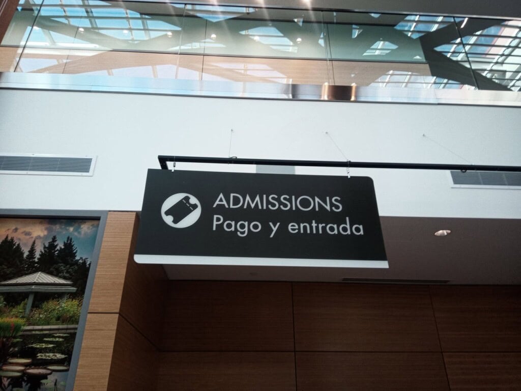 A black and white hanging sign reads ADMISSIONS Pago y entrada with an icon of a ticket. The sign is indoors, and above it is a glass ceiling with structural beams.