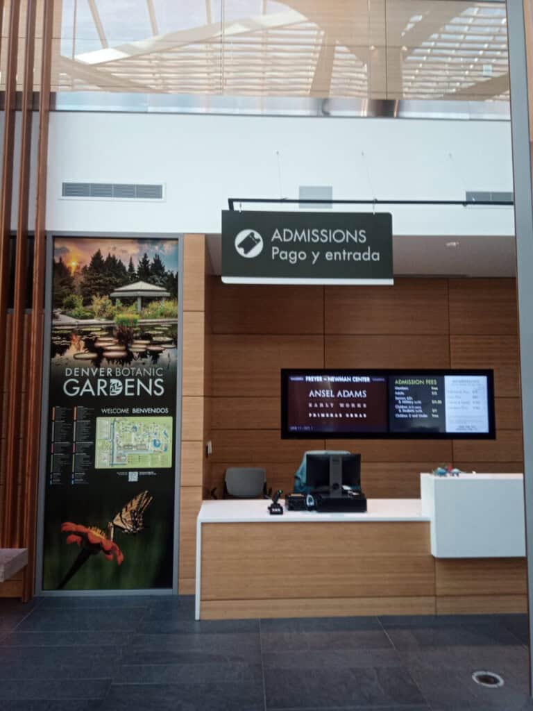 Admission desk at Denver Botanic Gardens. A digital sign above reads Admissions Pago y entrada. Informational posters and screens display ticket prices, a map, and Ansel Adams exhibit details. Modern, clean interior.