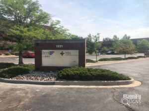 A brick sign in a parking lot displays 1221, ISYS MerIT, Harvey & Company Certified Public Accountants, and the Magnify Signs logo. Trees, parked cars, and a building are visible in the background.