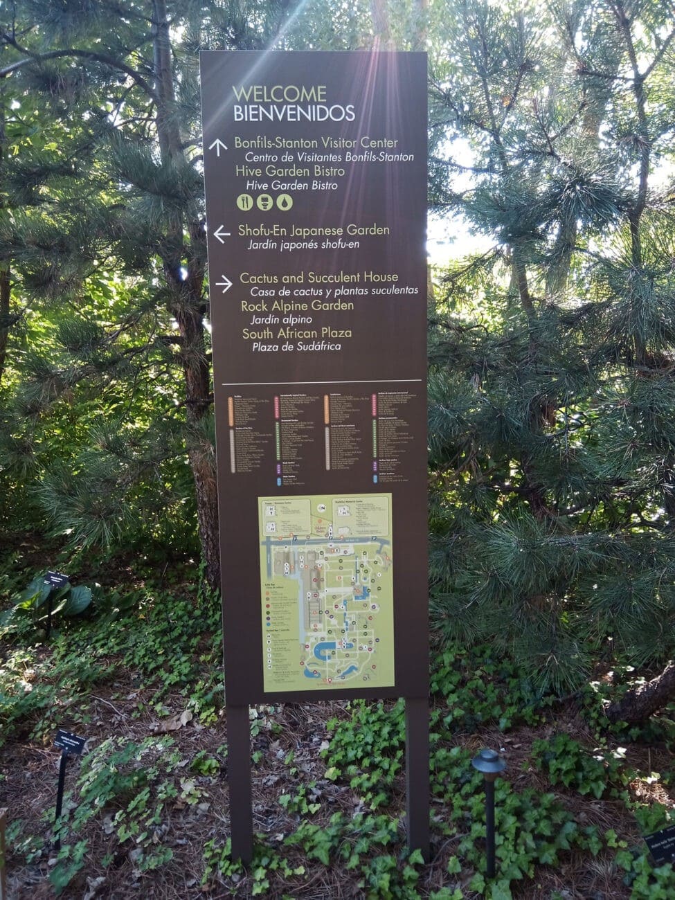 Tall brown sign in a garden with trees and greenery, displaying a welcome message in English and Spanish, directions to garden areas, a map, and a list of locations within the botanical garden.