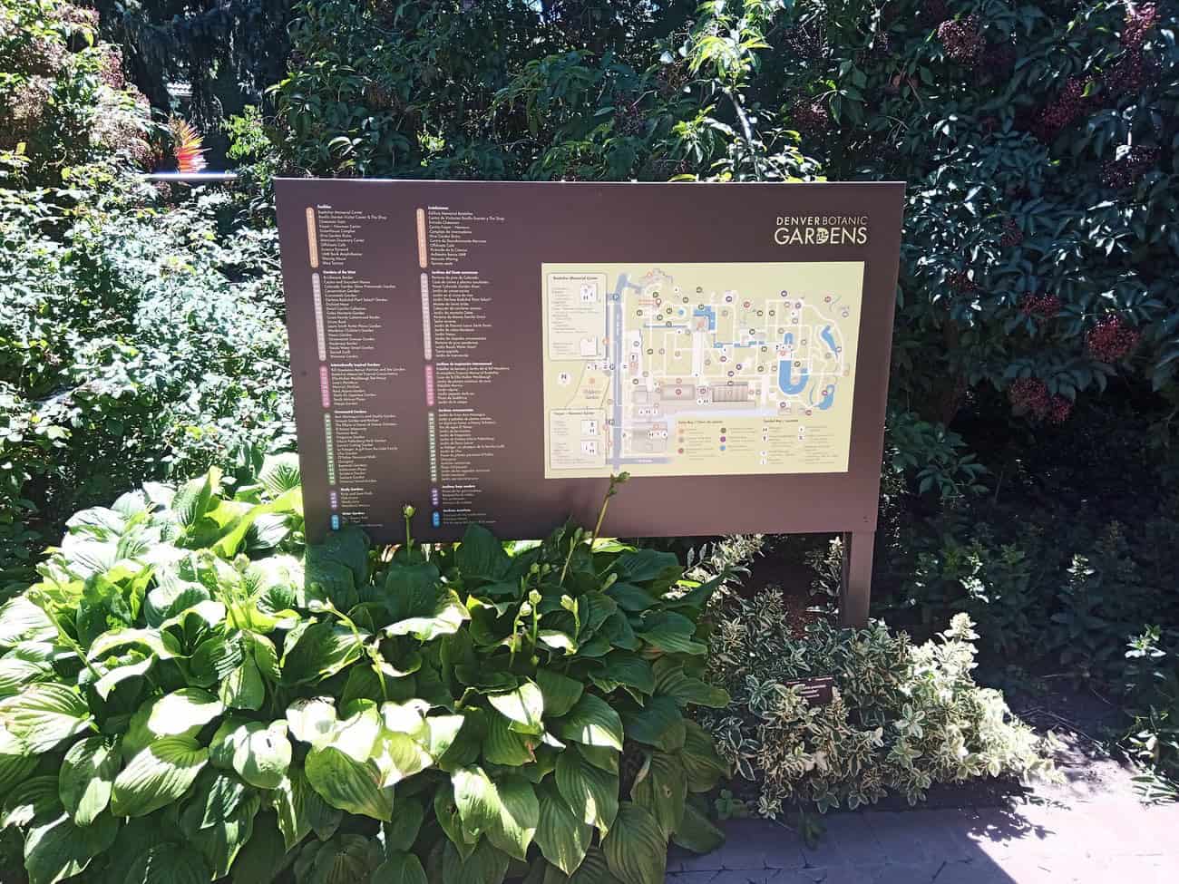 A large informational sign with a map and directory for Denver Botanic Gardens stands outdoors, partially shaded by surrounding green plants and trees. Sunlight brightens part of the sign and nearby foliage.