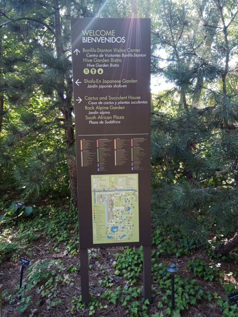 A tall, bilingual information sign in a botanical garden stands among green trees and shrubs. The sign has a map, garden locations, and directions in both English and Spanish. Sunlight filters through the branches above.