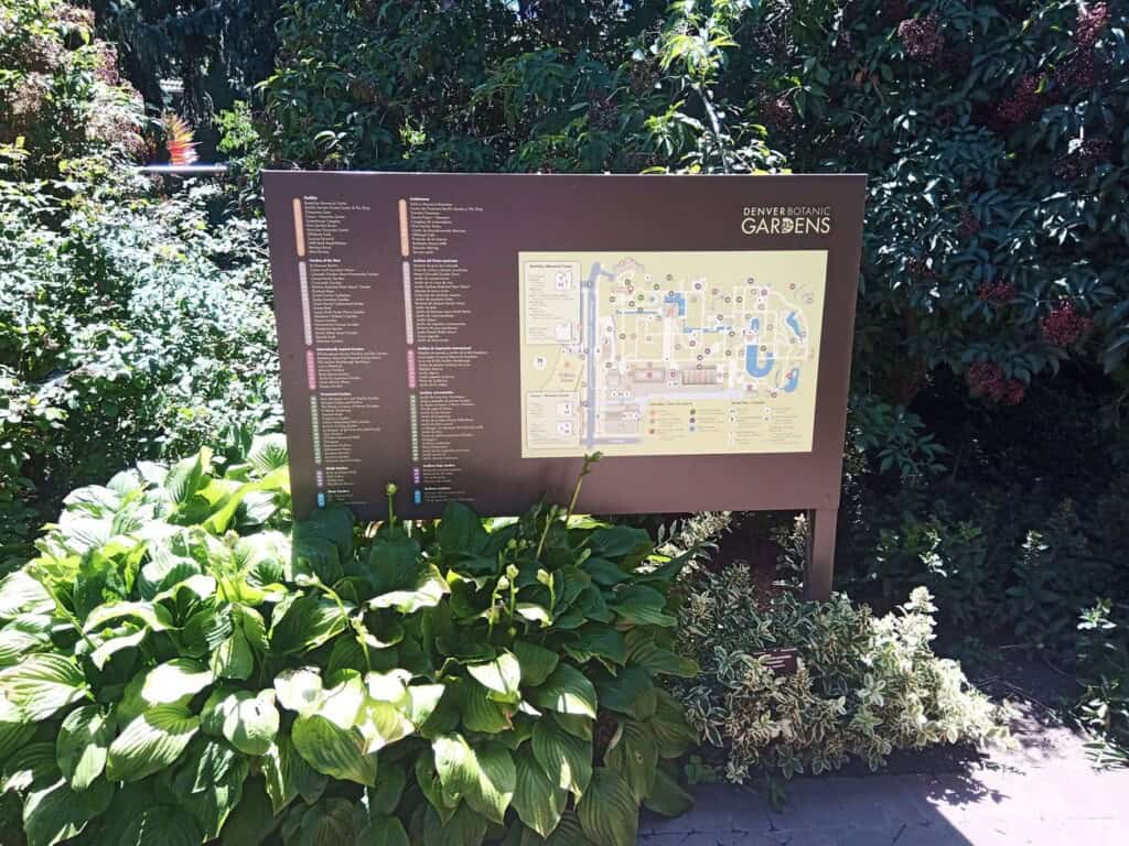 A large information sign with a map and directory for Denver Botanic Gardens stands amid lush green plants and foliage in a sunny outdoor setting.