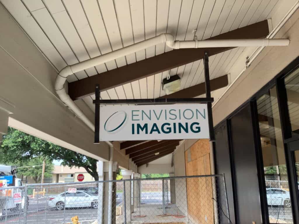 A hanging sign under a covered walkway reads Envision Imaging. There are construction barriers and fencing in front, and a street with parked cars is visible in the background.