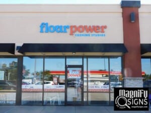 Storefront of Flour Power Cooking Studios with glass windows and doors, black awning, and colorful signage. The reflection of cars and trees is visible in the windows. A Magnify Signs logo appears in the bottom right corner.