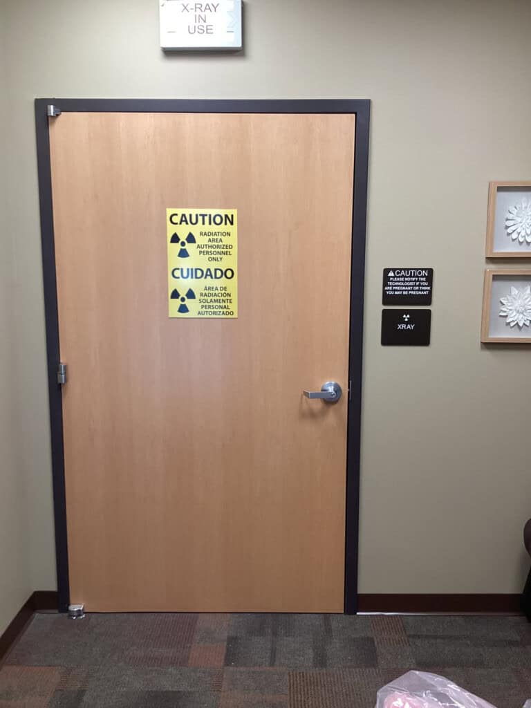 A closed wooden door with caution signs warning about radiation in English and Spanish. A black sign beside the door says X-RAY. The door is in a medical office with neutral-colored walls and carpet.