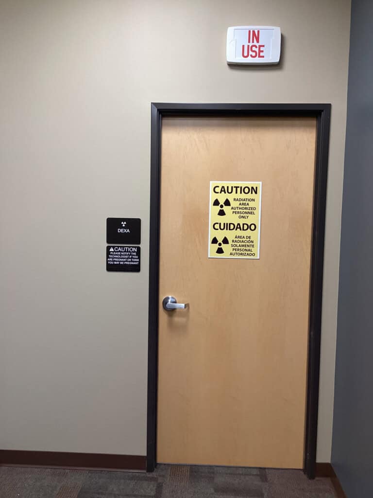 A closed wooden door with radiation warning signs in English and Spanish. Above the door is a lit IN USE sign. To the left, a wall plaque reads DECA and a caution message. The surrounding walls are beige.