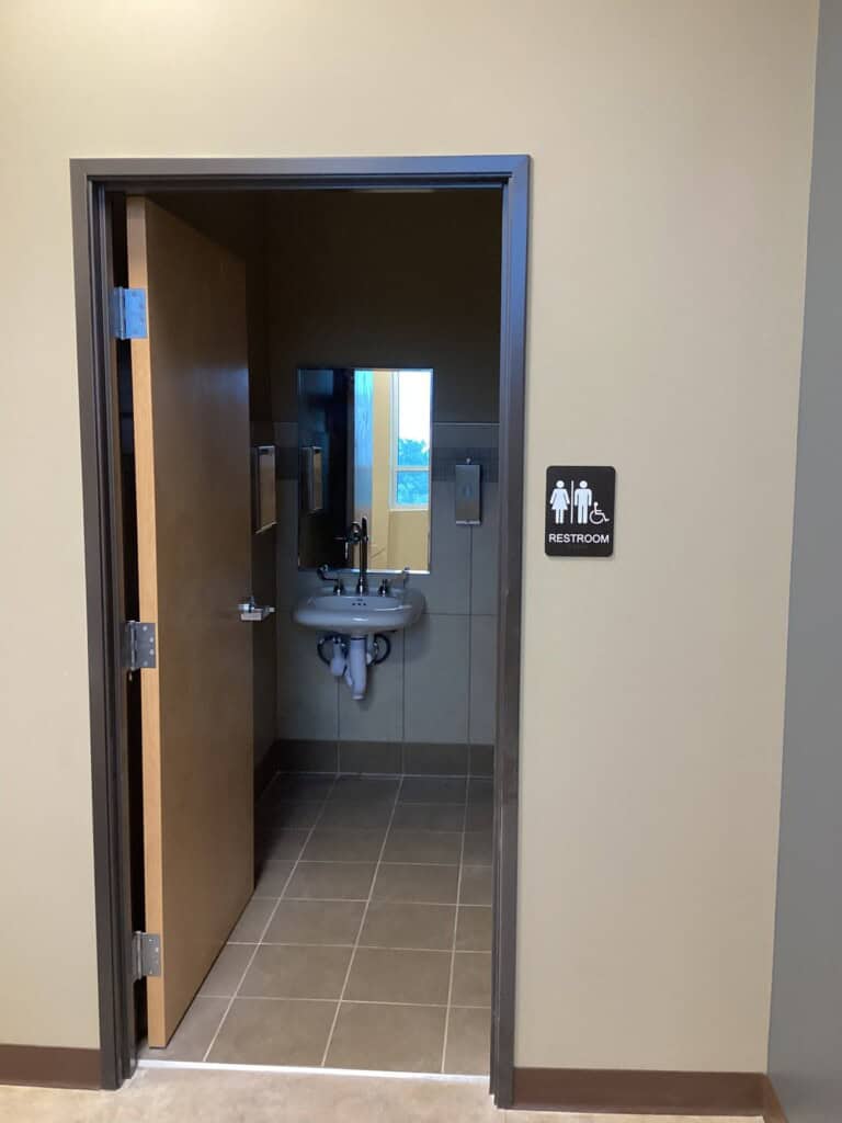 A restroom with an open door shows a sink, a mirror, and a soap dispenser inside. A sign beside the door indicates it is a gender-neutral and accessible restroom.
