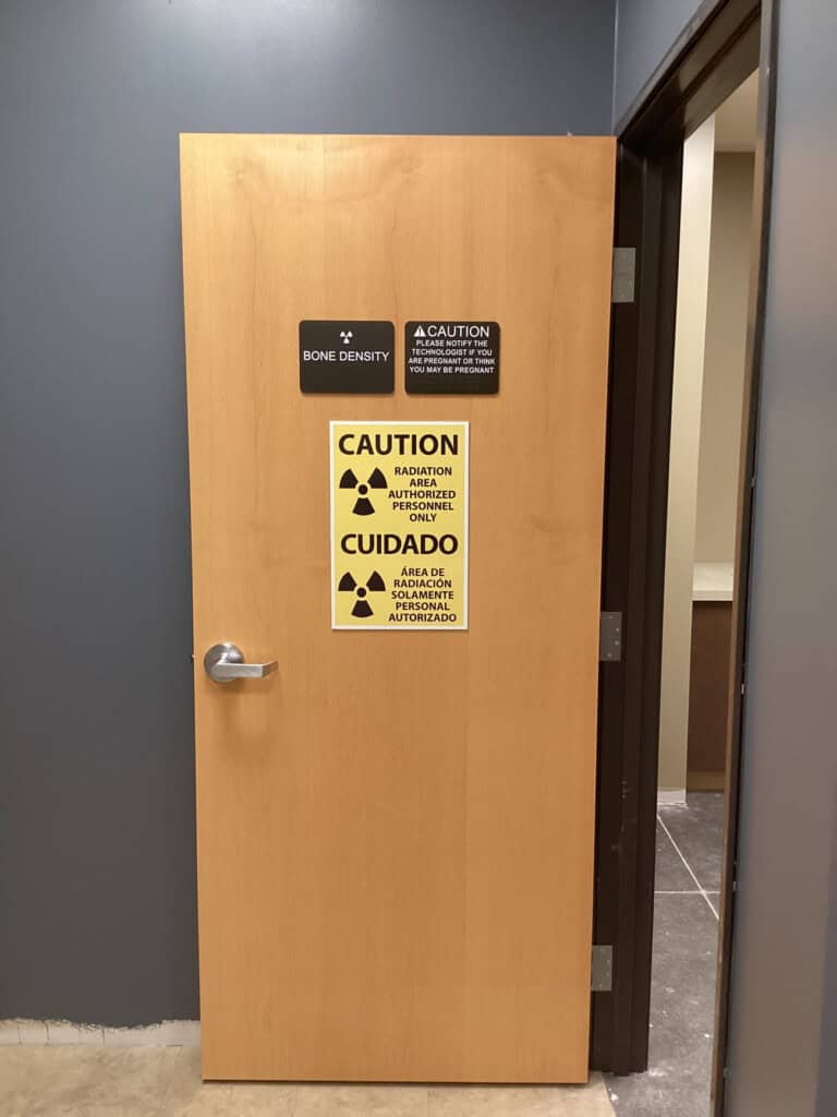 A wooden door with warning signs, including a radiation symbol and text in English and Spanish indicating Bone Density and Caution: Radiation Area. Authorized Personnel Only. The door is slightly open in a medical facility.