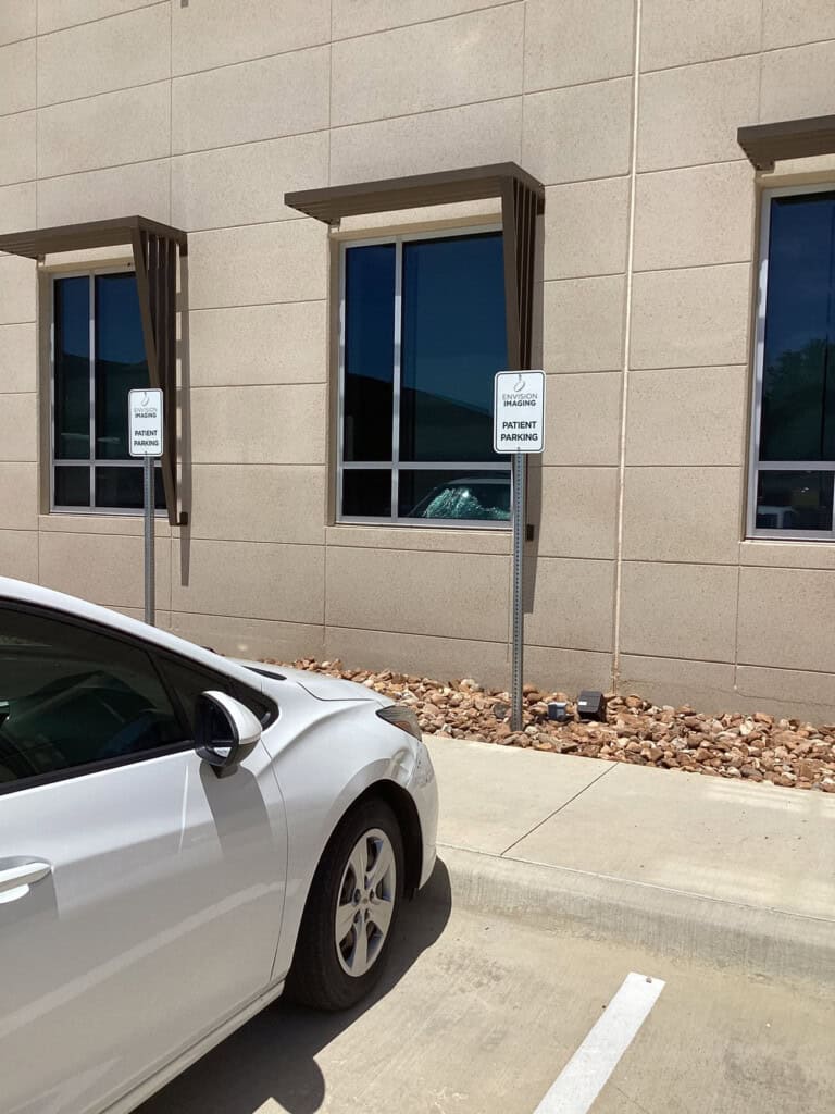 A white car is parked in front of a beige building, next to a sign that reads Volunteer Patient Parking. The area has rocky landscaping and two windows are visible on the building.