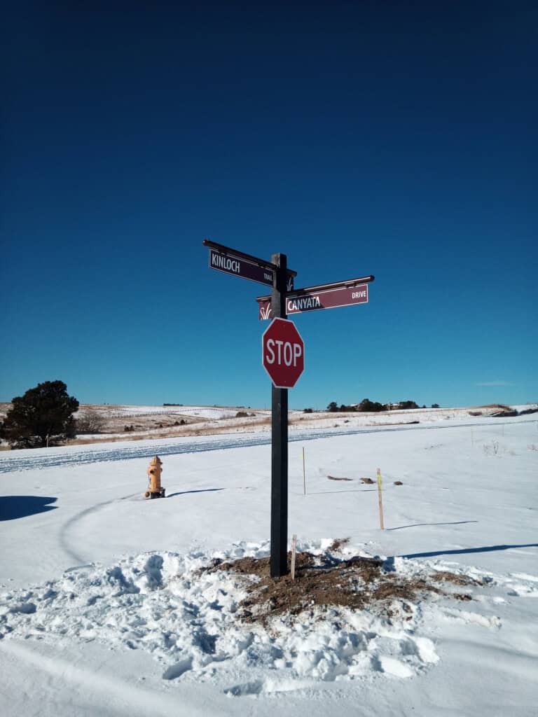 A stop sign stands at a snowy intersection with street signs for Kinloch Drive and Canyata Drive under a clear blue sky. A yellow fire hydrant and distant trees are visible in the background.