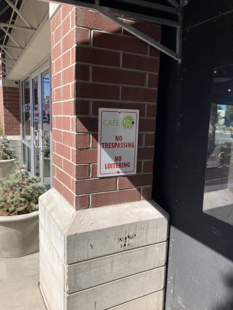 A sign on a brick pillar outside a building reads CAFE 18, No Trespassing, No Loitering. Nearby are large windows and plants in concrete planters. Sunlight casts shadows on the sidewalk.