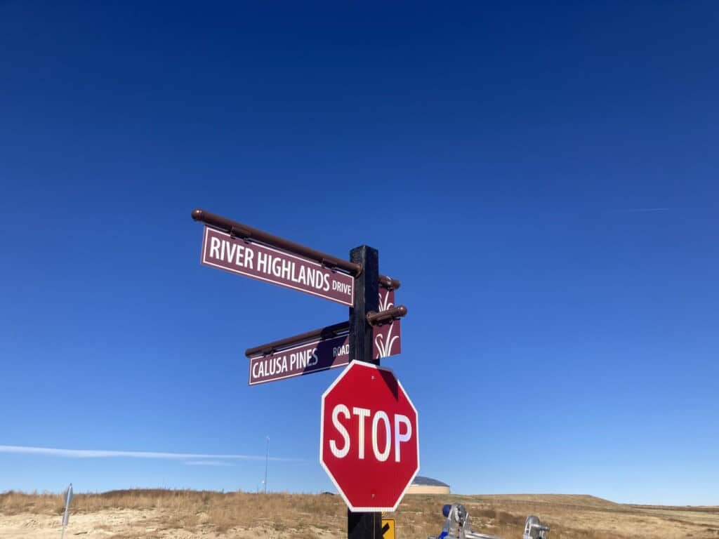 A stop sign stands at an intersection with street signs for River Highlands Drive and Calusa Pines Drive against a clear blue sky and open landscape.