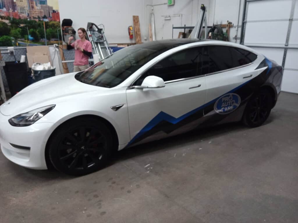 A white Tesla sedan with a blue and black mountain-themed graphic and Pro Auto Care logo is parked inside a garage. A person in a pink shirt stands in the background.