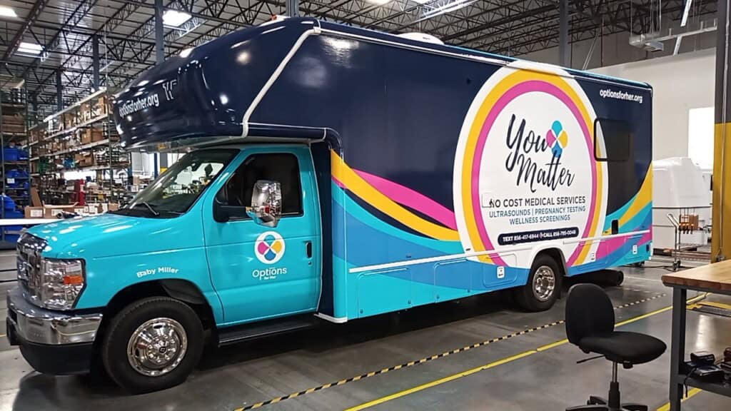 A brightly colored mobile clinic van with the words You Matter and No Cost Medical Services on its side is parked inside a warehouse. The van is blue with multicolored accents and the Options logo.