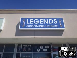 A storefront sign reads Legends Grooming Lounge in white letters on a blue background, with barber pole graphics on each side. The Magnify Signs logo appears in the bottom right corner.