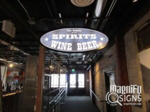 An oval ceiling sign reads SPIRITS WINE BEER in large letters, hanging in an industrial-style hallway with exposed pipes and dim lighting. The Magnify Signs logo appears in the bottom right corner.