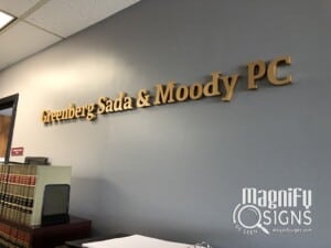 Wall with gold 3D letters spelling Greenberg Sada & Moody PC above an office desk, shelves with legal books, and a logo for Magnify Signs in the lower corner.