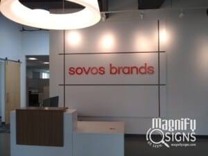 A modern office reception area with a white wall featuring the red text “sovos brands.” The Magnify Signs logo is visible in the bottom right corner of the image.
