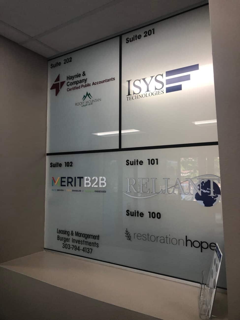 Window panels display office suite numbers and business names, including Haynie & Company, ISYS Technologies, VERITB2B, Reliant, Restoration Hope, and a leasing/investment contact number, viewed from a hallway.