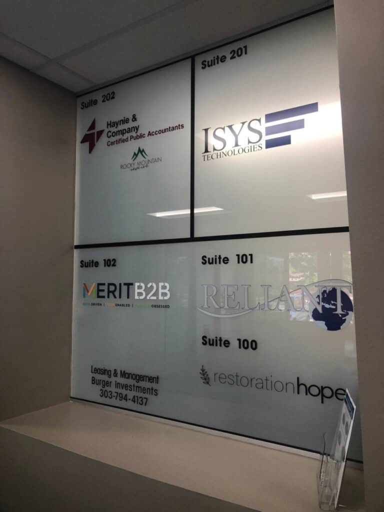 A glass wall displays six business signs with suite numbers, including accounting, technology, marketing, realty, leasing, and a nonprofit organization. Sunlight filters through the windows behind the signs.