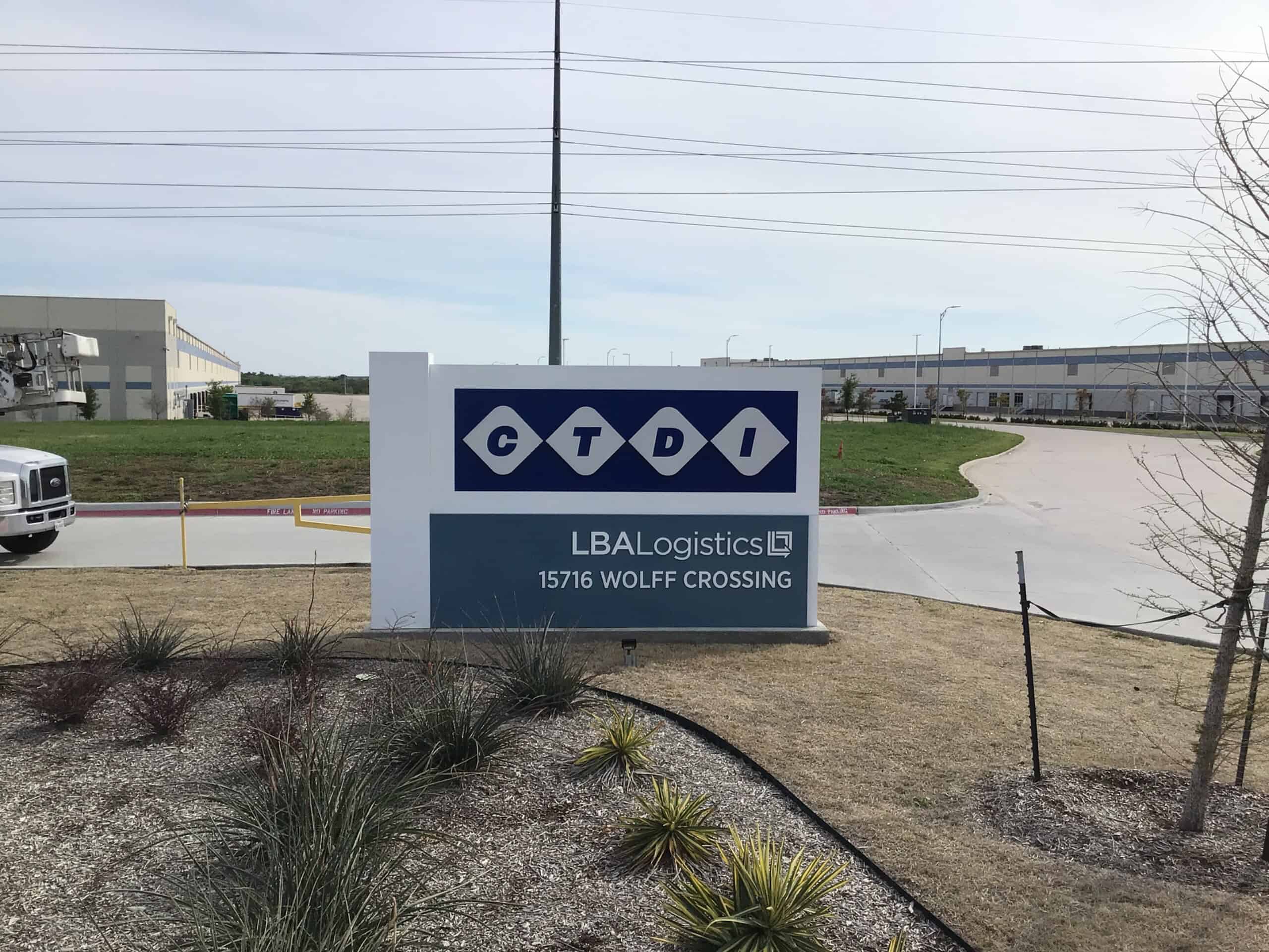 A sign in front of a business park reads CTDI in blue diamonds and LBA Logistics, 15716 Wolff Crossing. Industrial buildings and a truck are visible in the background.