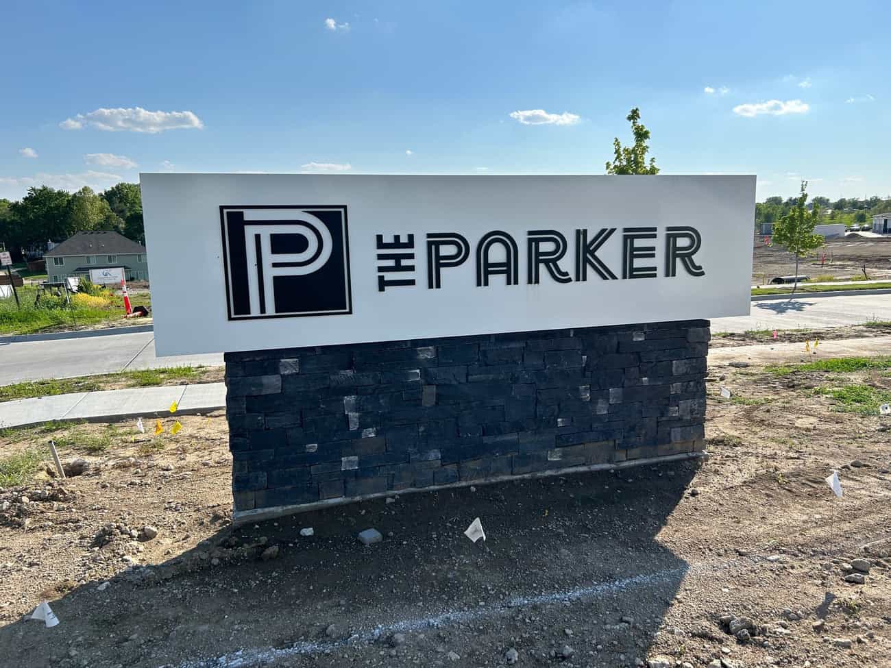 A white sign with black text that reads THE PARKER and a large stylized P stands on a stone base at the edge of a construction site, with houses and trees visible in the background.