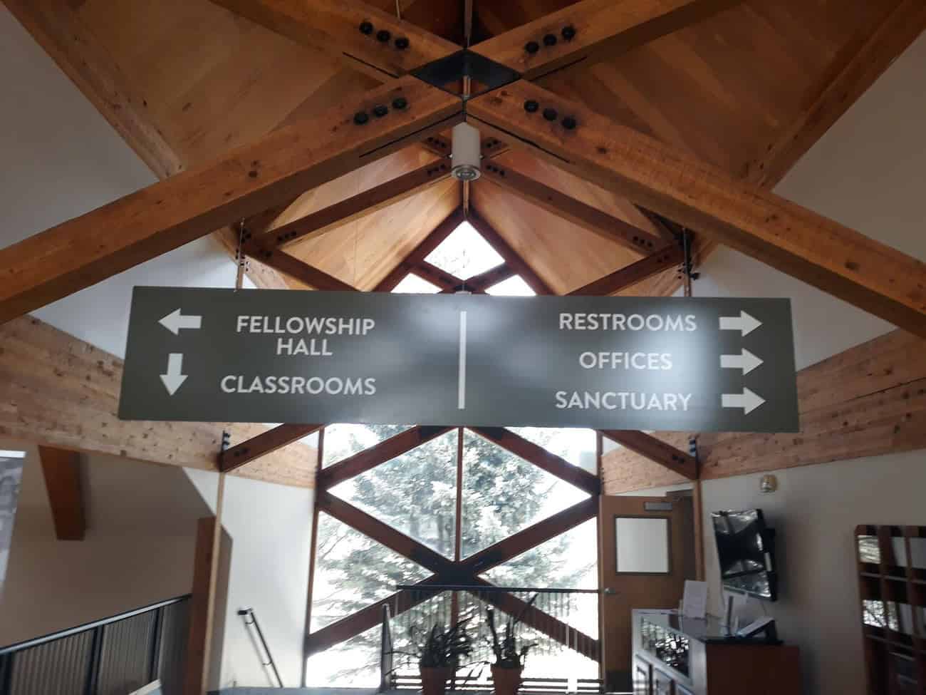 A large directional sign hangs from a wooden-beamed ceiling. It points left to Fellowship Hall and Classrooms, and right to Restrooms, Offices, and Sanctuary. Sunlight streams through large windows behind.
