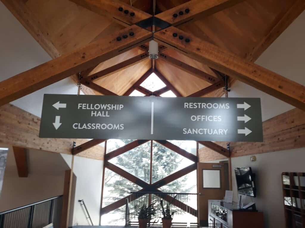 A large indoor sign with arrows directs left to Fellowship Hall and Classrooms, and right to Restrooms, Offices, and Sanctuary. The sign hangs from a wooden ceiling with exposed beams and large windows in the background.