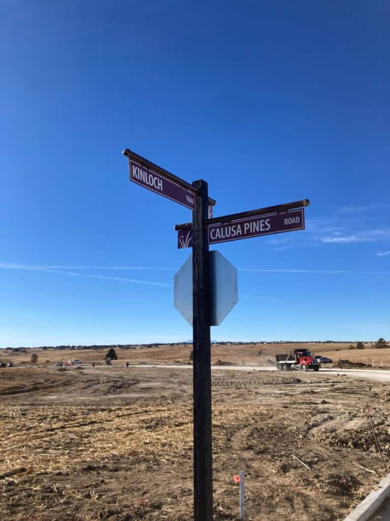 A street sign at an intersection shows Kinloch Trail and Calusa Pines Road against a clear blue sky. In the background, there is empty land and construction equipment.