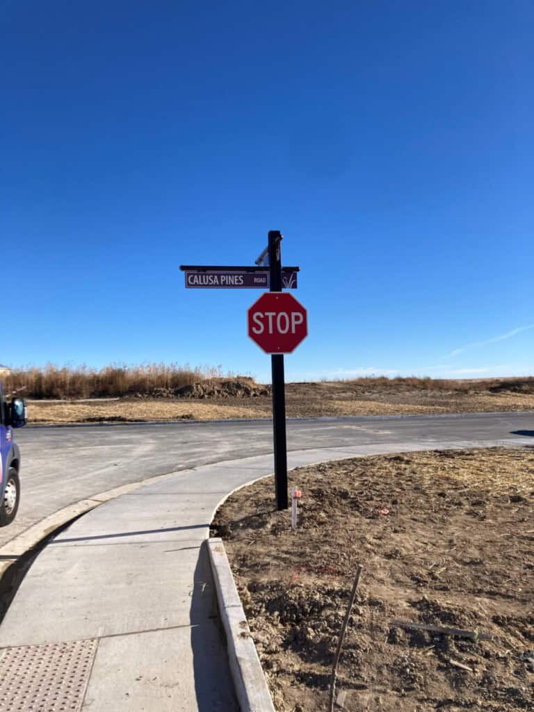 A stop sign and a street sign for Calusa Pines Road stand at the edge of a curved road under a clear blue sky, with dry grass and dirt in the background.