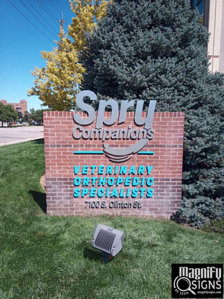 A brick sign reads Spry Companions VETERINARY ORTHOPEDIC SPECIALISTS, 7100 S. Clinton St. with trees and grass around it. A Magnify Signs logo is in the bottom right corner.