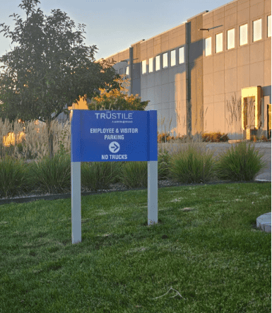 A blue sign on a grassy area reads TruStile Employee & Visitor Parking. No Trucks in front of a modern gray building with large windows and landscaped shrubs.