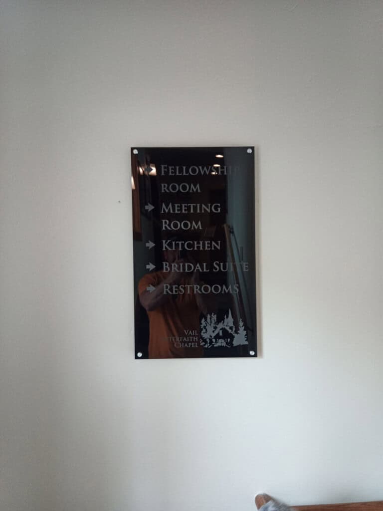 A wall-mounted sign with arrows directs to the Fellowship Room, Meeting Room, Kitchen, Bridal Suite, and Restrooms at Vail Interfaith Chapel. A person’s reflection is faintly visible in the sign.
