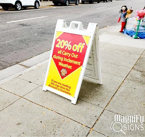 A sidewalk sign reads “20% off all Carry Out during Inclement Weather. If you are reading this sign, then the discount is available.” It stands on a city sidewalk near a street, with flowers and a doll in the background.