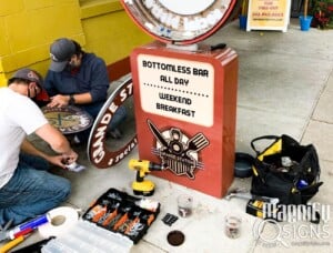 Two workers in masks install or repair a vintage-style sign outside a restaurant. Tools, screws, and a yellow drill are spread on the sidewalk. The sign advertises a bottomless bar and weekend breakfast.