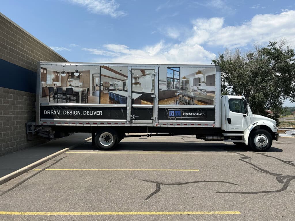 A white delivery truck parked beside a building, featuring large advertisements on its side displaying kitchen and bathroom designs with the text DREAM. DESIGN. DELIVER and the BKC Kitchen and Bath logo.