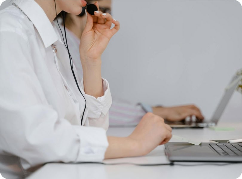 Two people working at a desk; the person in the foreground is wearing a headset microphone and using a laptop, suggesting a customer service or call center setting. The background is out of focus.