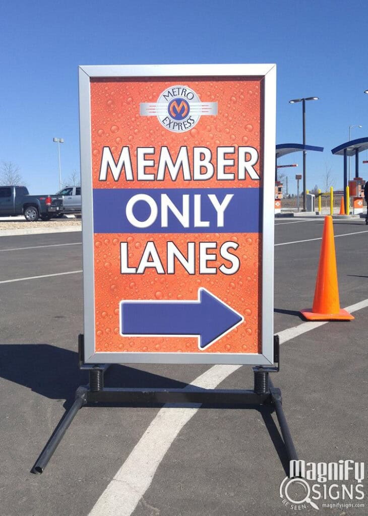 A roadside sign reads Metro Express Member Only Lanes with a right-pointing arrow, placed on an asphalt surface near traffic cones and a toll booth under a clear blue sky.