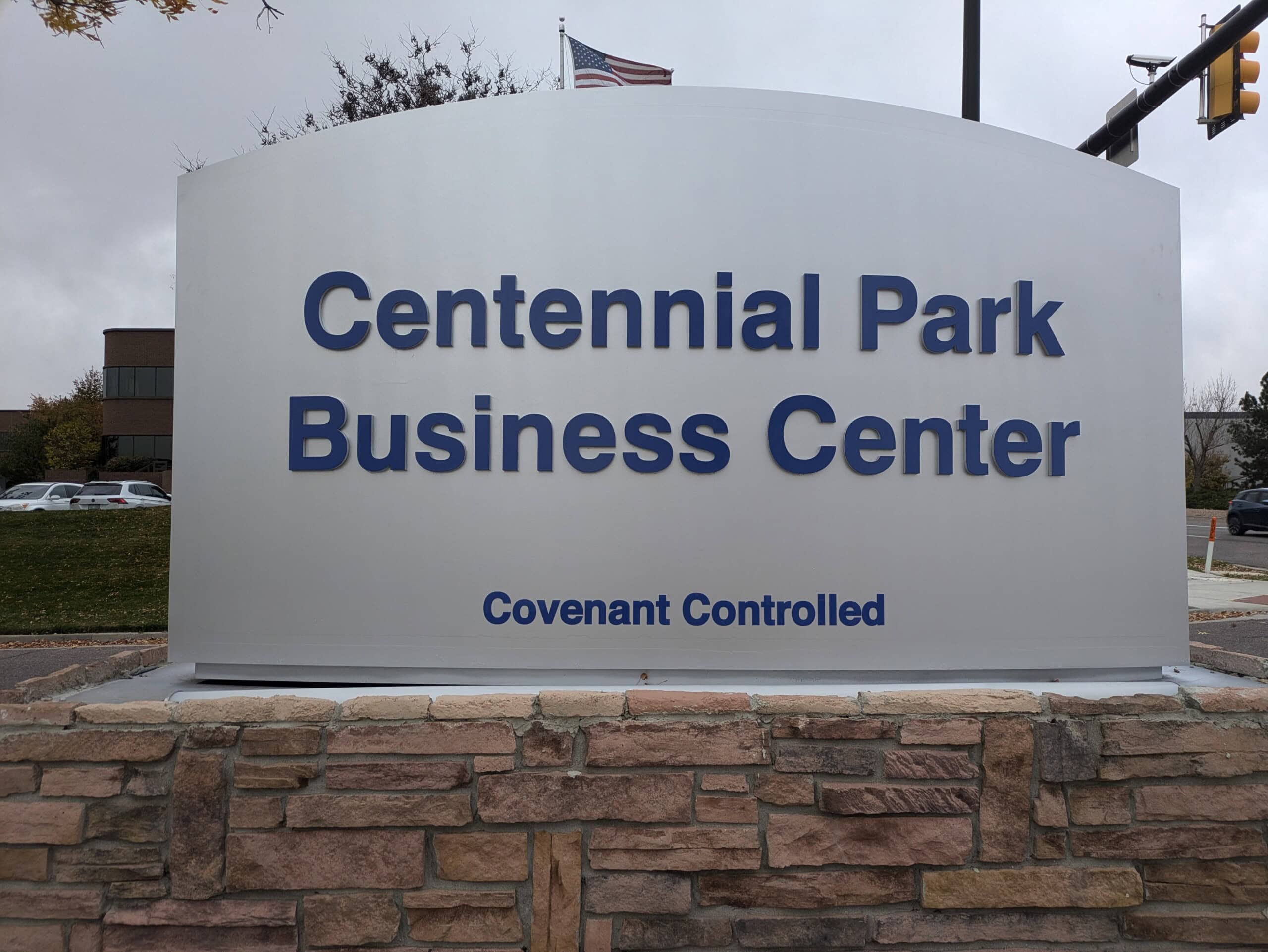 A large outdoor sign reads Centennial Park Business Center with Covenant Controlled below it. The sign is set on a stone base, and there is a grassy area, trees, and a building in the background.