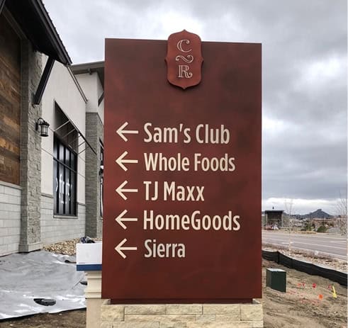 A large outdoor sign with five arrows pointing left, listing Sam’s Club, Whole Foods, TJ Maxx, HomeGoods, and Sierra against a cloudy sky background.